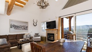 Een ruime woonkamer met een open haard, een grote televisie en een eethoek met uitzicht op de bergen.