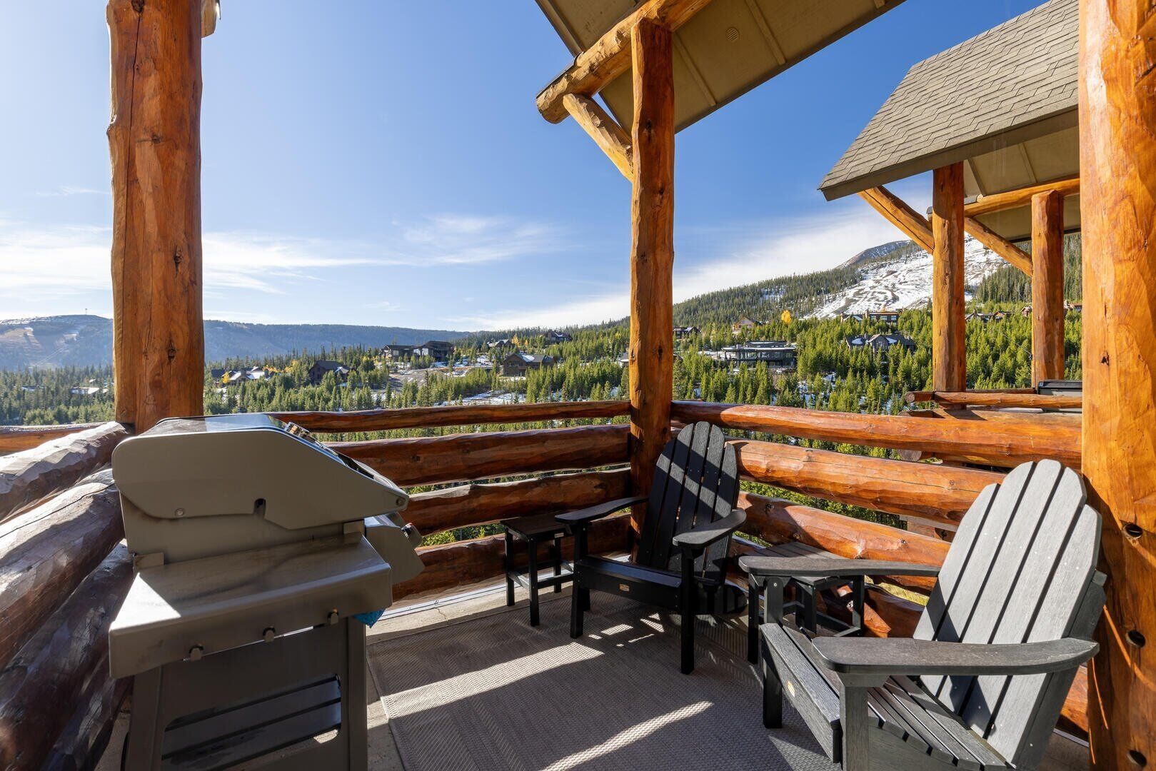 Una terraza de madera con una parrilla, sillas y vistas a un paisaje montañoso nevado.