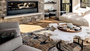 A modern living room with a stone fireplace, a mounted TV, and a large window overlooking mountains. - 7 Deep Forest 6 Bedroom Home (Big Sky)