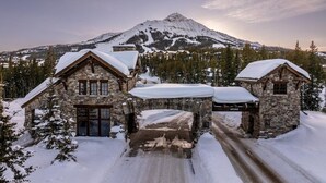 A stone lodge with a snow-covered entrance and a mountain backdrop. - 7 Deep Forest 6 Bedroom Home (Big Sky)
