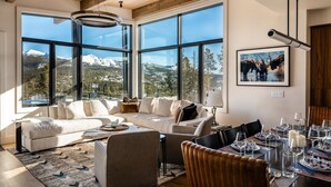 A spacious living area with a large window overlooking a mountainous landscape, a cozy seating arrangement, and a dining table set for a meal. - 7 Deep Forest 6 Bedroom Home (Big Sky)