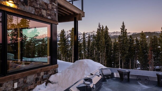 A modern outdoor hot tub area with a stone wall and glass windows overlooking a snowy mountain landscape. - 7 Deep Forest 6 Bedroom Home (Big Sky)