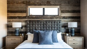 A bedroom with a wooden headboard, a large window, two bedside tables with lamps, a bed with blue bedding, and a nightstand with drawers. - 7 Deep Forest 6 Bedroom Home (Big Sky)