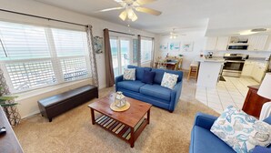 A living room with a blue sofa, a wooden coffee table, and a bench. There is a kitchen area with white cabinets and a microwave.