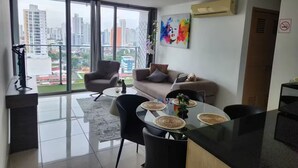 Ein modernes Wohnzimmer mit einer Couch, Sesseln, einem Esstisch mit Platzsets und einem Blick auf die Stadt durch große Fenster.