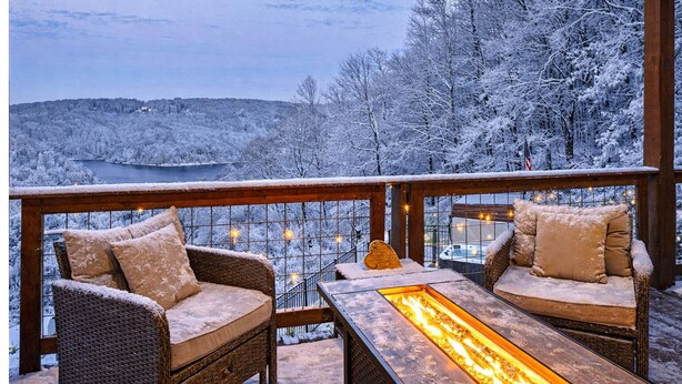 Un balcon doté de meubles en osier et d’une table à plateau de verre intégrant un foyer, offrant une vue sur un paysage enneigé et une étendue d’eau.