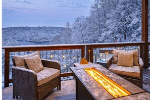 Un balcón con muebles de mimbre y una mesa con superficie de vidrio que incorpora un elemento decorativo de fuego, con vistas a un paisaje nevado y una masa de agua.