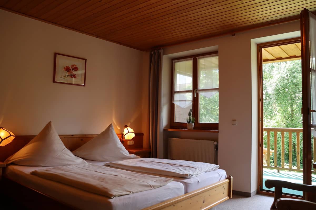 A double bed with two pillows, a bedside table with a lamp, a window with a view of greenery, and a wooden ceiling.