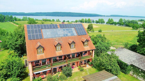 Un edificio de dos pisos con techo de tejas rojas y paneles solares. Cuenta con varios balcones y ofrece vistas a un lago y colinas boscosas al fondo.