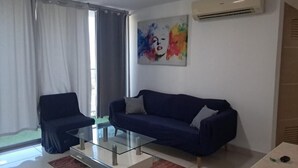 Una sala de estar con un sofá azul oscuro, una mesa de centro de vidrio y una silla. Hay un cuadro abstracto y colorido en la pared y un equipo de aire acondicionado.