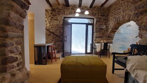 Quarto rústico com paredes de pedra aparente, soalho de madeira, cama com colcha verde, escrivaninha com cadeira e vista através de uma porta.