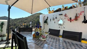 Um pátio externo com mesa, cadeiras e guarda-sol. Há plantas em vasos e objetos decorativos na parede.