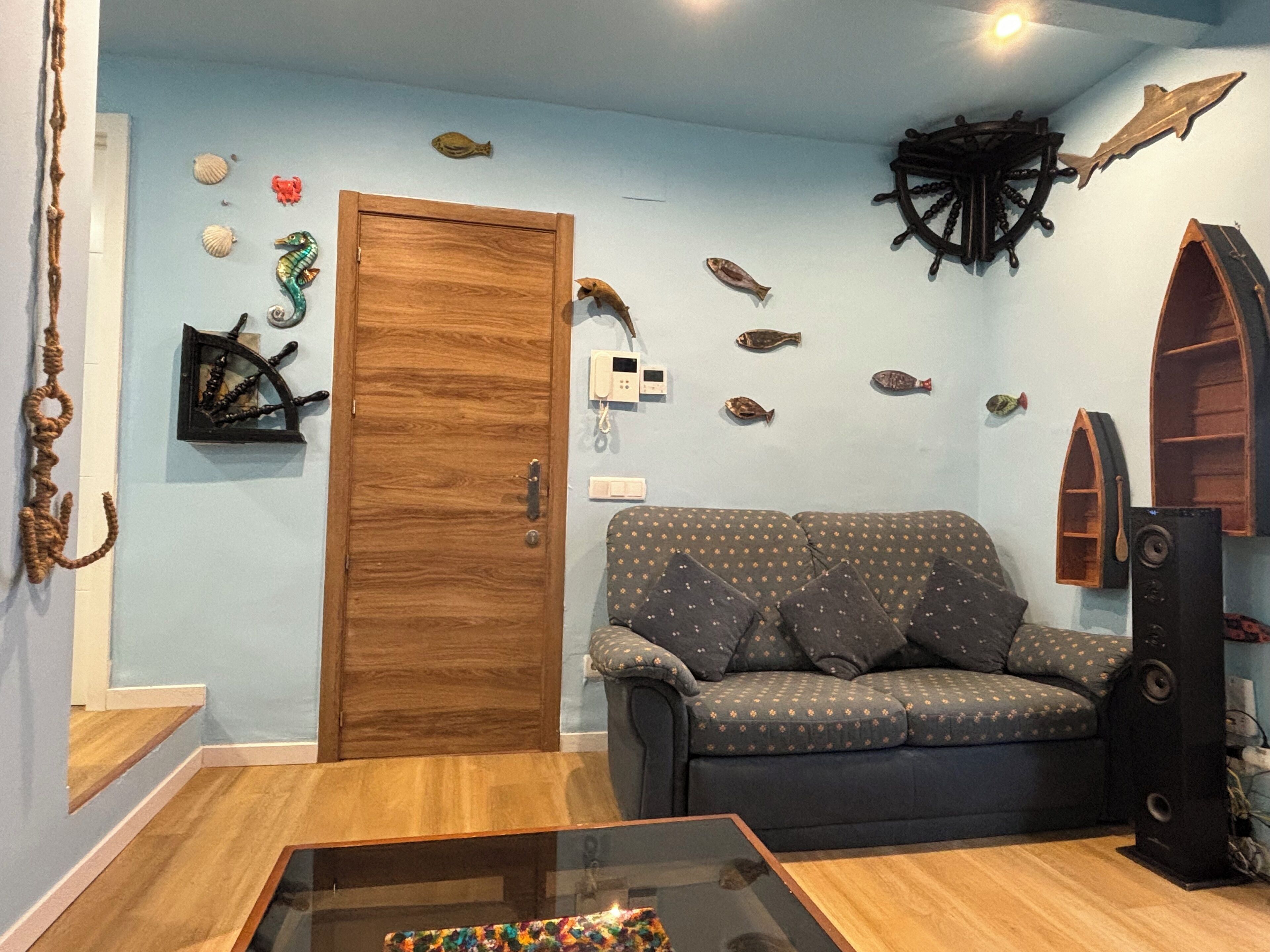 Un salon avec une porte en bois, un canapé à motifs, une table basse en verre et des poissons décoratifs accrochés au mur.
