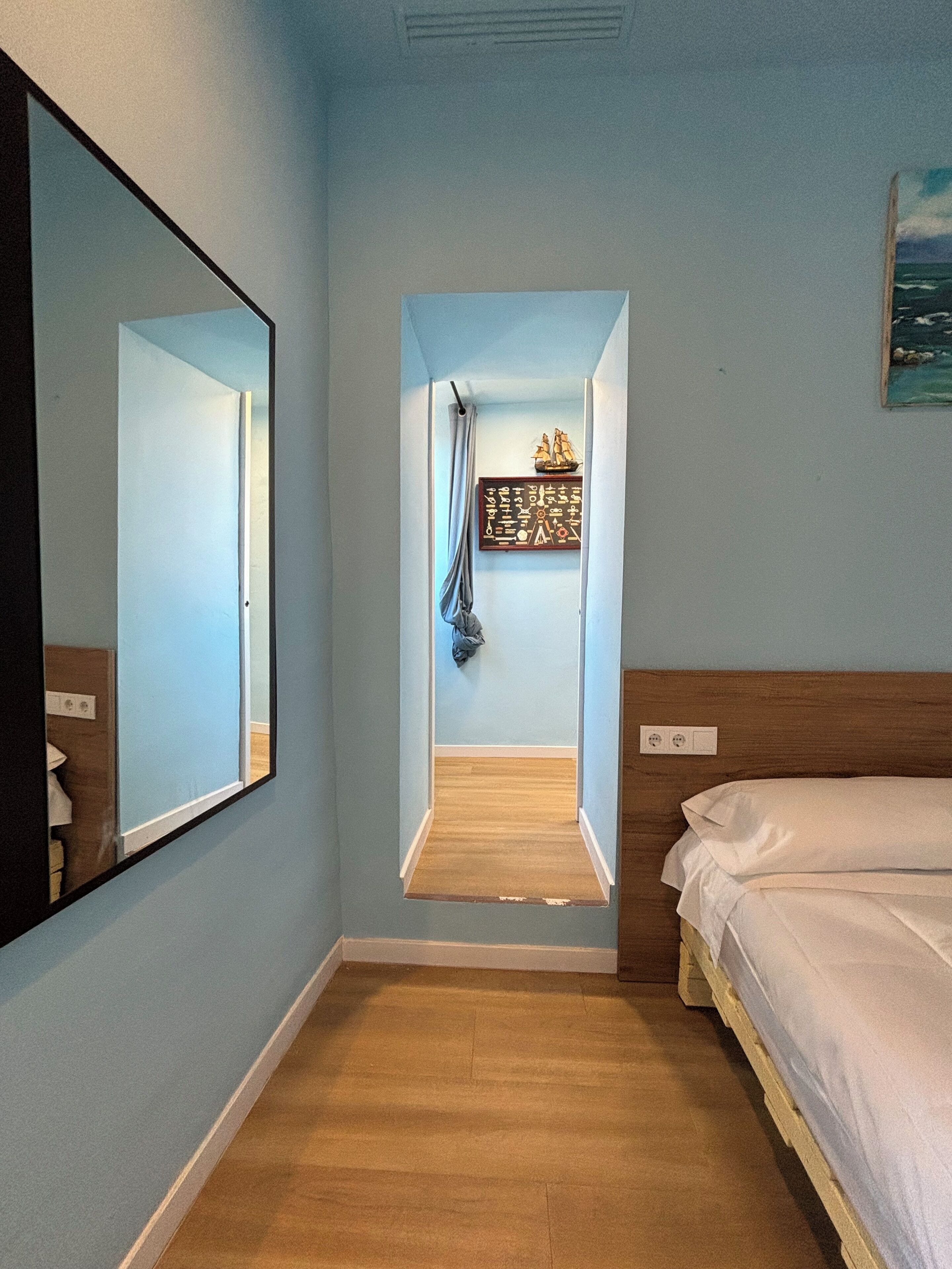 Ein Zimmer mit einem Bett, einem Spiegel, einem kleinen Fenster mit Vorhang und einer Wanddekoration.