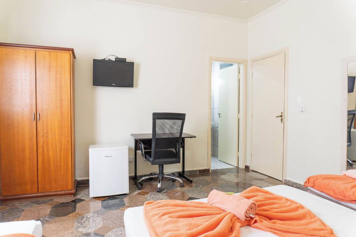 Ein Hotelzimmer mit Kleiderschrank, Schreibtisch mit Stuhl, kleinem Kühlschrank und zwei Betten mit orangefarbenen Handtüchern.