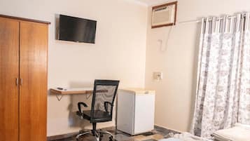 Um pequeno espaço de escritório com um guarda-roupa de madeira, uma escrivaninha com cadeira, um frigorífico branco pequeno e um ar condicionado fixado na parede, além de uma cortina.