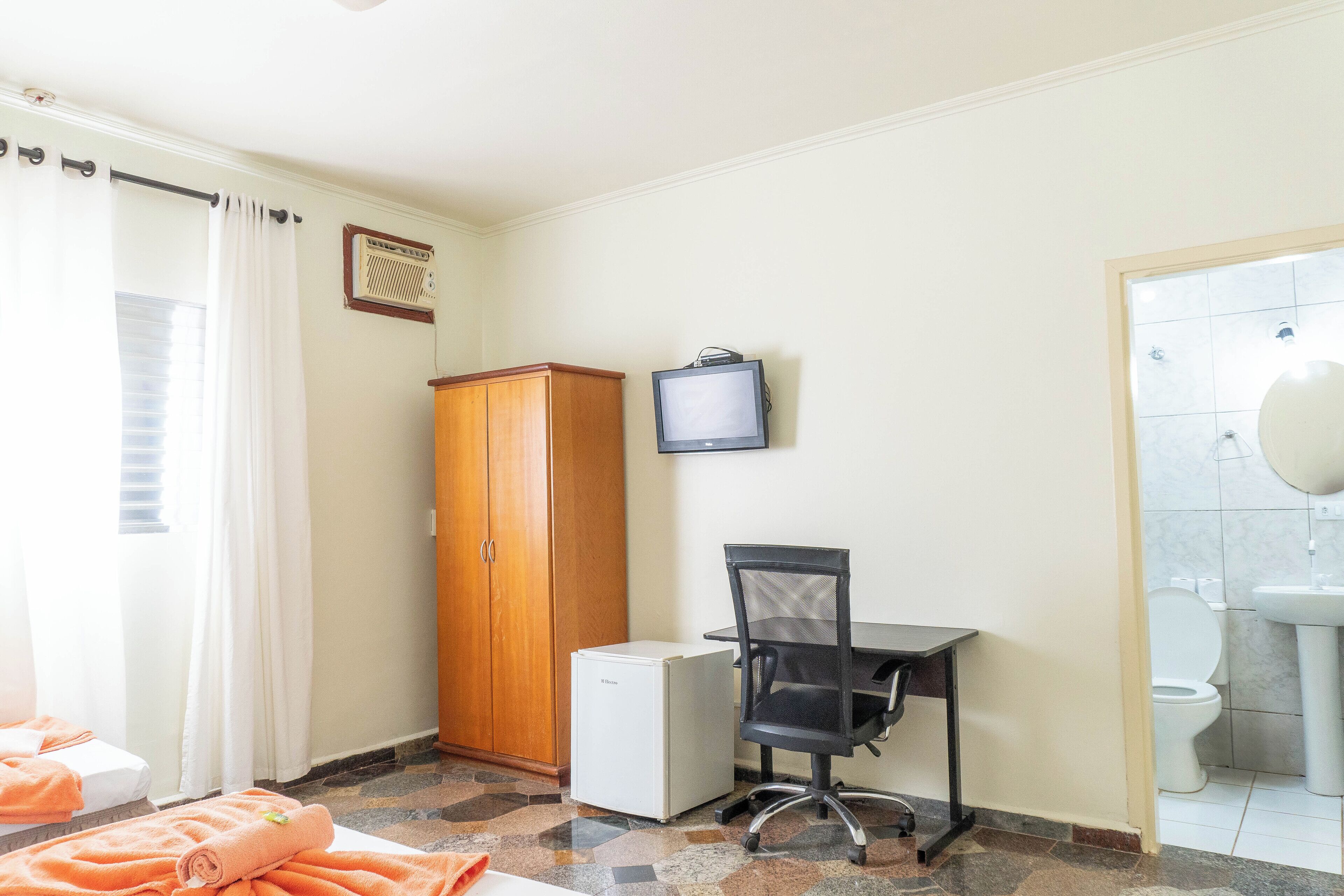 Una habitación con cama, escritorio y silla, armario, televisión y baño con lavamanos e inodoro.