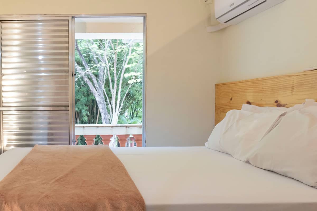 Ein Schlafzimmer mit einem Bett, einem hölzernen Kopfteil, einem Fenster mit Blick auf Bäume und einer Klimaanlage.