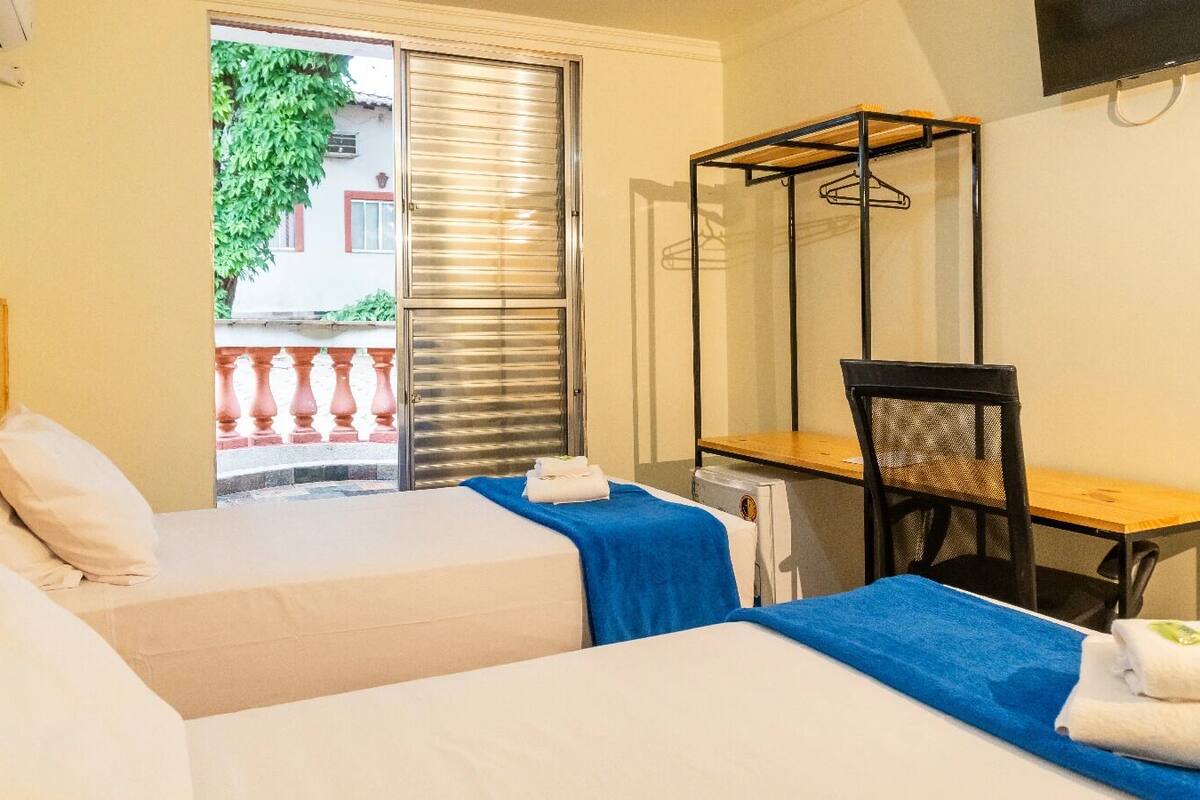Ein Hotelzimmer mit zwei Betten, einem Schreibtisch, einem Stuhl und einem Fenster mit Blick auf einen Balkon und ein Gebäude.