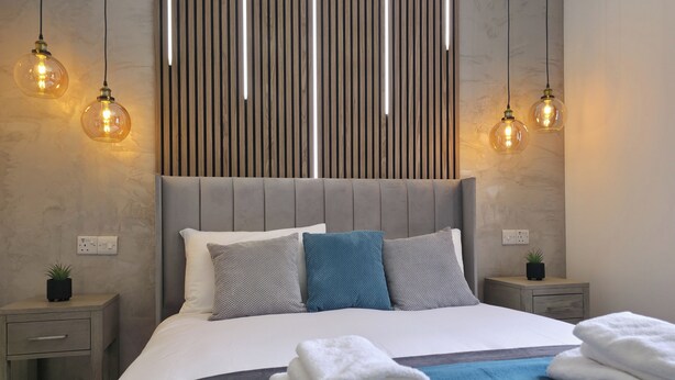 Ein modernes Schlafzimmer mit grauem gepolsterten Kopfteil, einem großen Bett mit blau-weißer Bettwäsche und zwei Nachttischen mit Lampen.