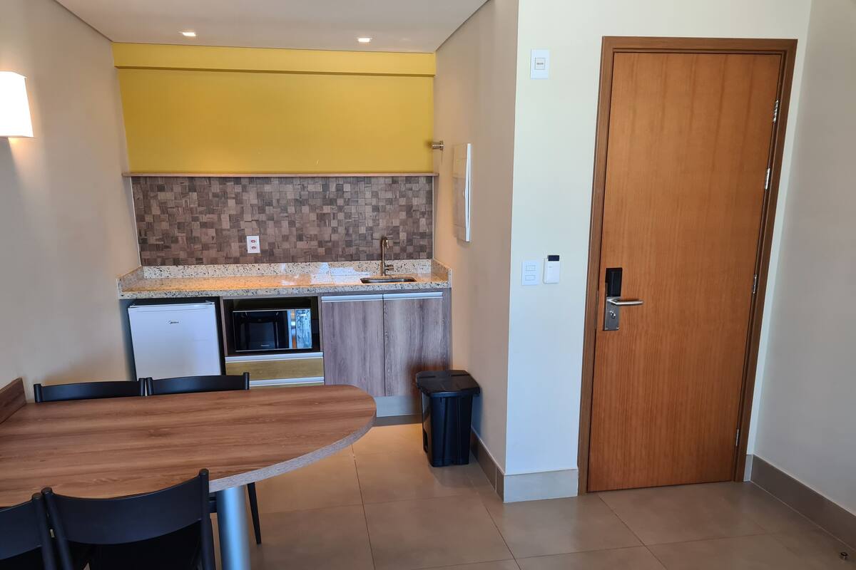 Cocina compacta con microondas, horno y lavamanos. Hay una mesa de comedor con sillas y un cubo de basura. Se ve una puerta de madera a la derecha.