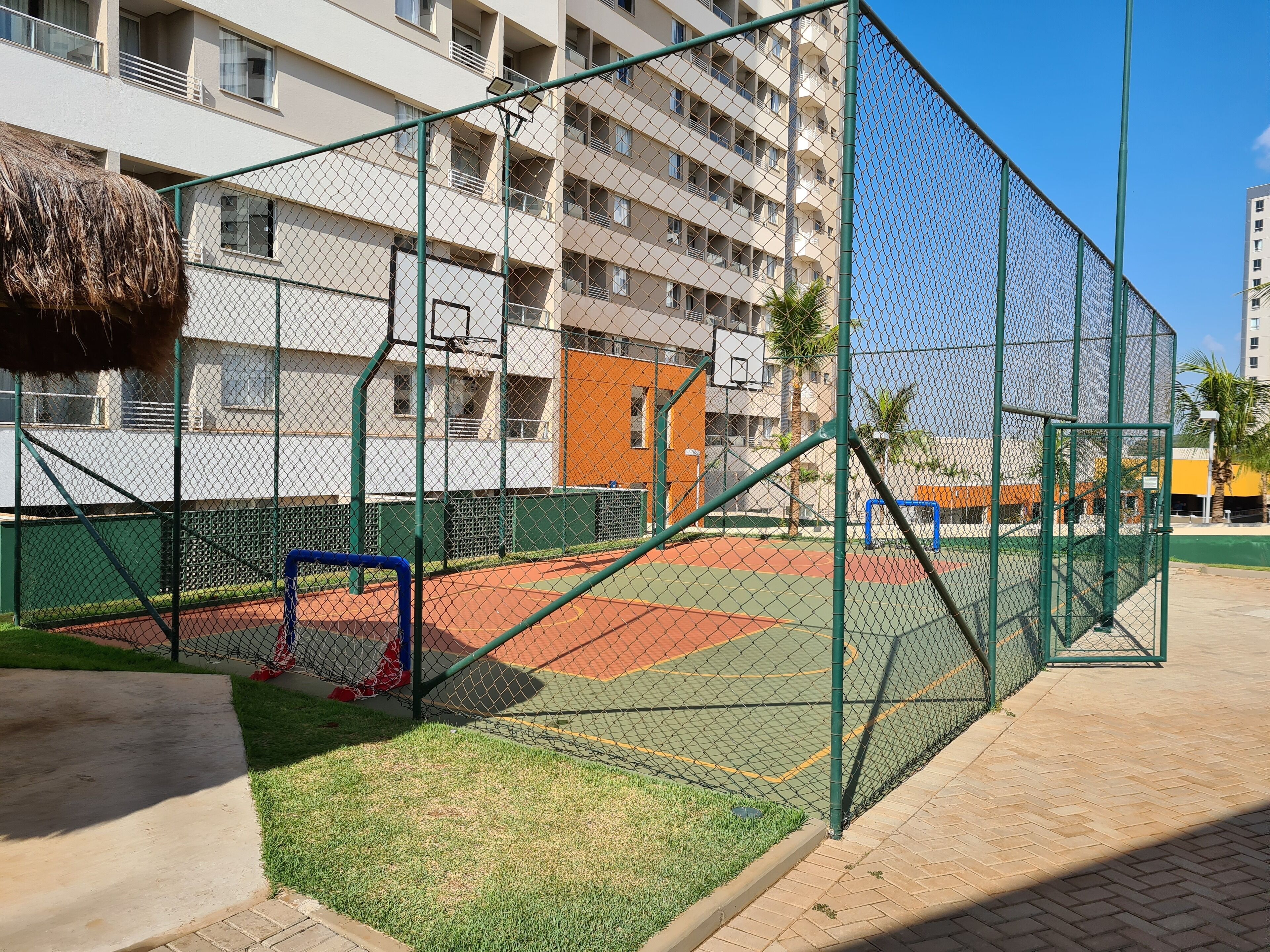 A fenced sports court with a basketball hoop, surrounded by a multi-story building with balconies.