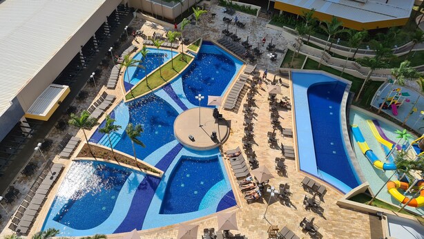 A large outdoor swimming pool with multiple sections and a central circular area, surrounded by lounge chairs and umbrellas. - Hospede-se no Solar das Águas (Olímpia)