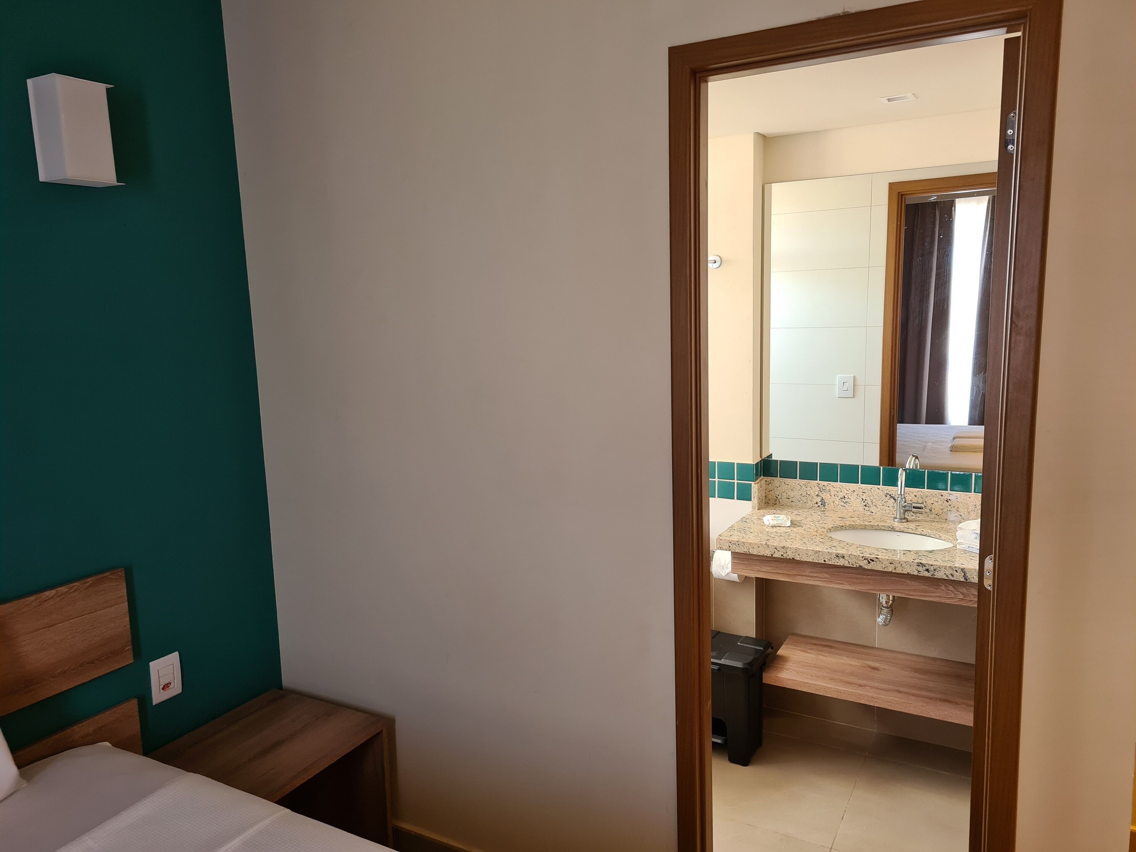 A hotel room with a bed, a small bedside table, a mirror, a bathroom with a sink and a toilet, and a trash bin.