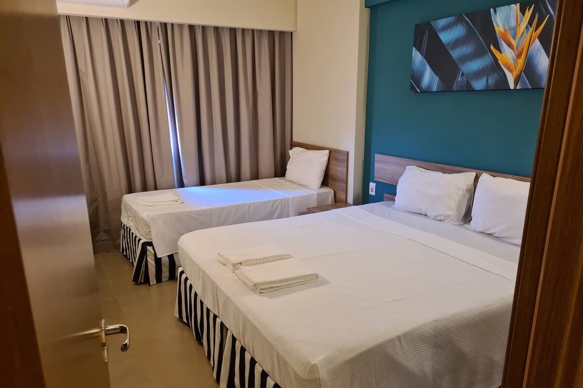 Habitación de hotel con dos camas, un ventanal con cortinas, una pared de acento color aguamarina y un cuadro sobre las camas.