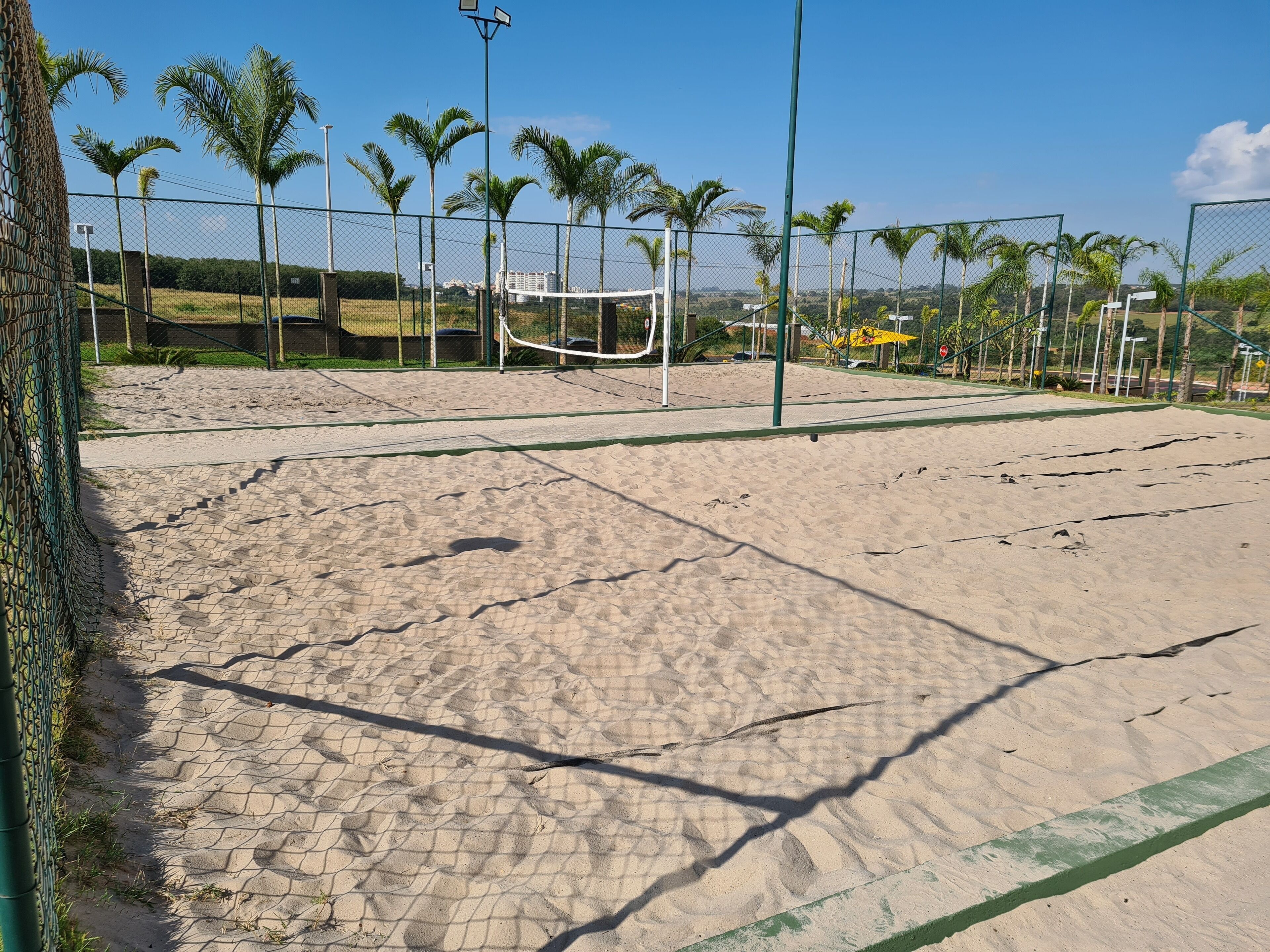 A sandy volleyball court with a net, surrounded by a chain-link fence and palm trees.