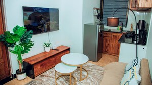 A compact living space with a TV mounted on the wall, a wooden bench, a small dining table, a sofa, and a kitchen area with a refrigerator and stove.
