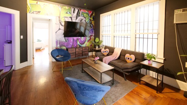 A modern living room with a sofa, coffee table, and blue chairs. A TV is mounted on the wall, and there is a window with blinds. - Miami style gem/central location (Miami)