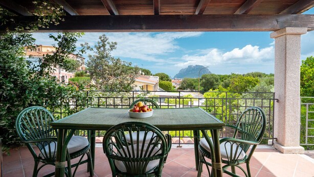A covered patio with a table and chairs, a bowl of fruit, and a view of buildings and trees.