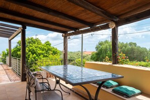 A covered patio with a table and chairs, a railing, and a view of greenery and buildings.