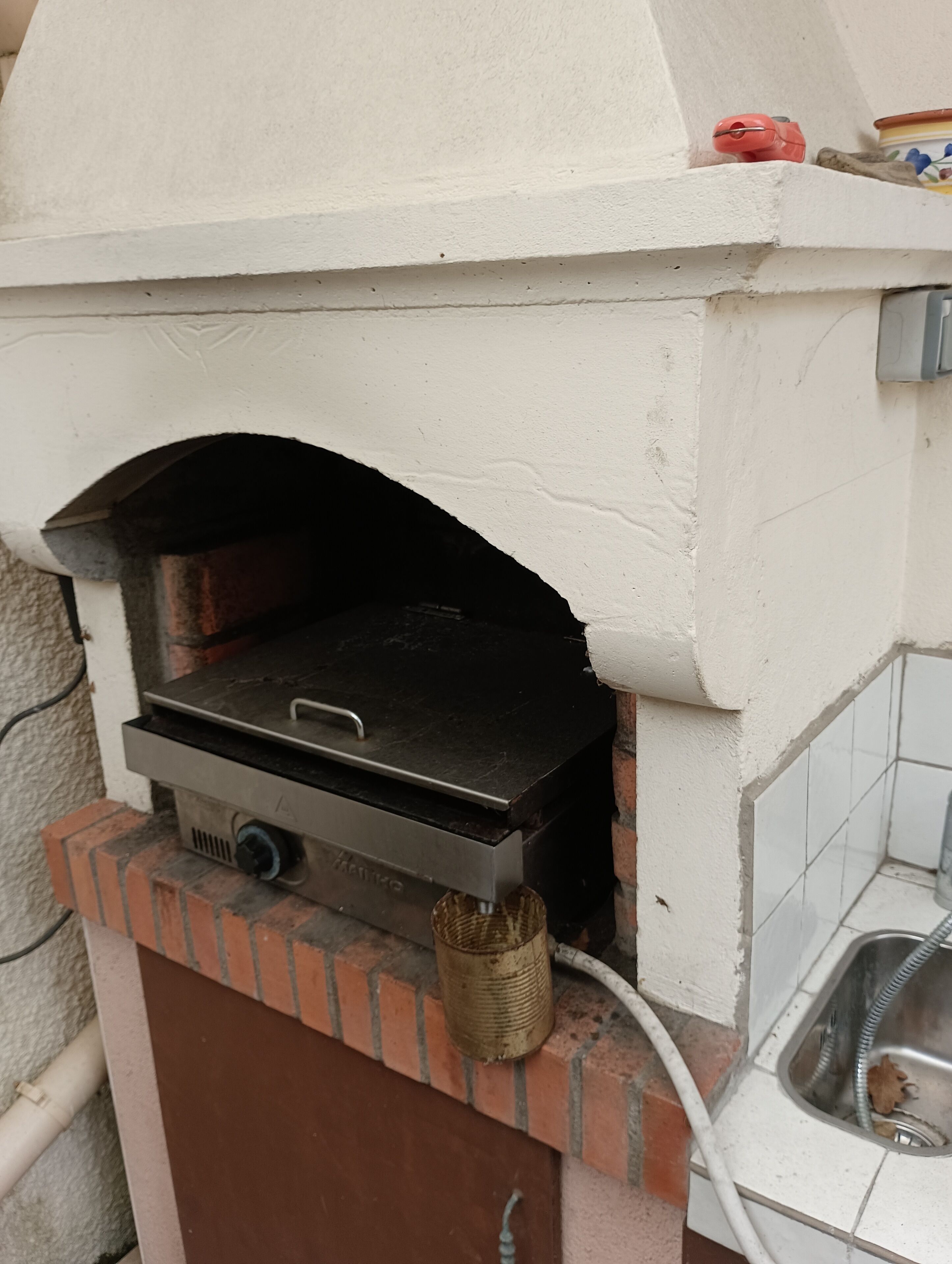 A brick-built outdoor oven with a metal door, a control knob, and a hose connected to it.