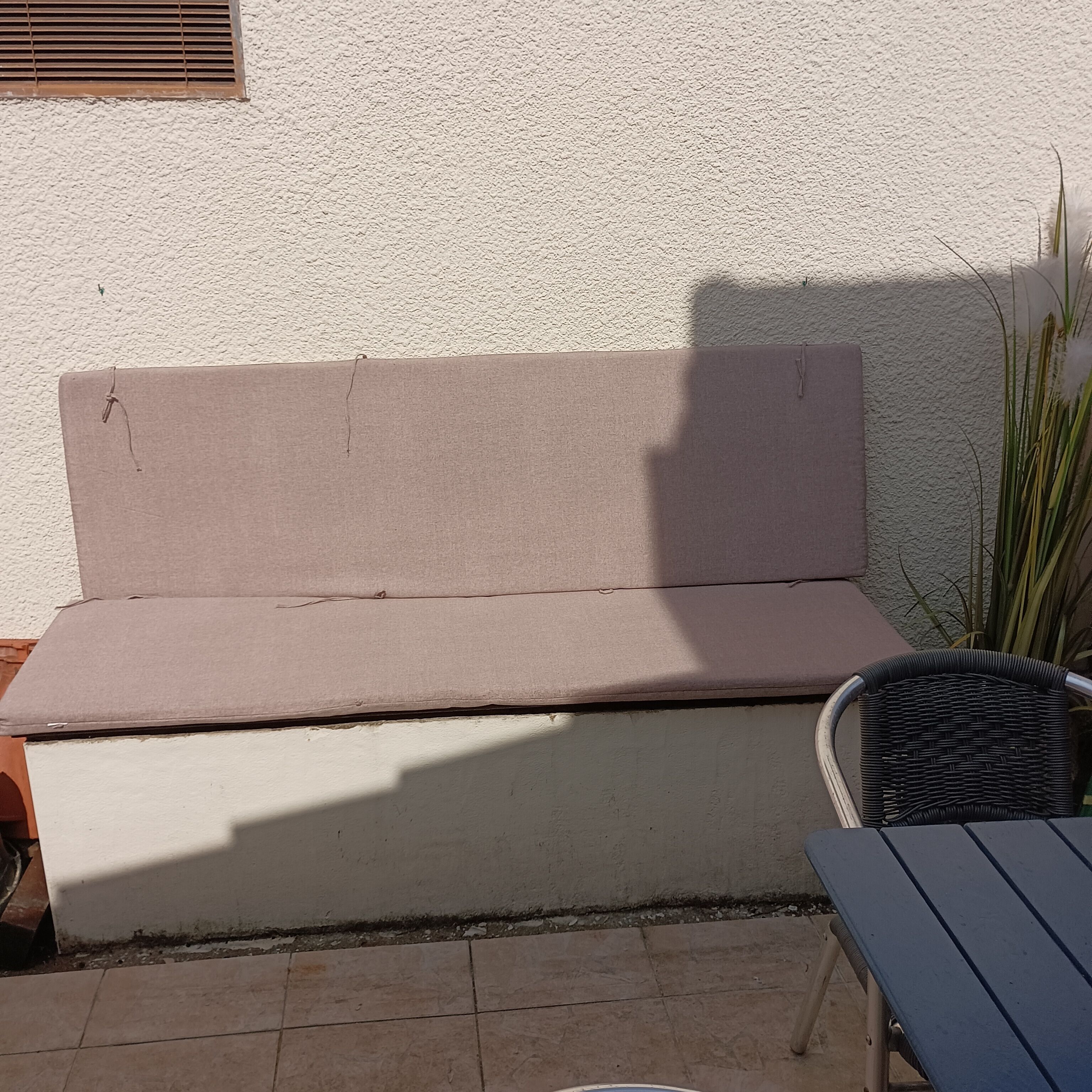 A beige outdoor bench against a textured wall, with a wicker chair and a potted plant nearby.
