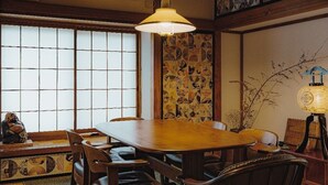Une salle à manger de style japonais traditionnel, avec une grande table en bois, des chaises en bois foncé, une lampe suspendue et un mur en pierre orné d’œuvres d’art.