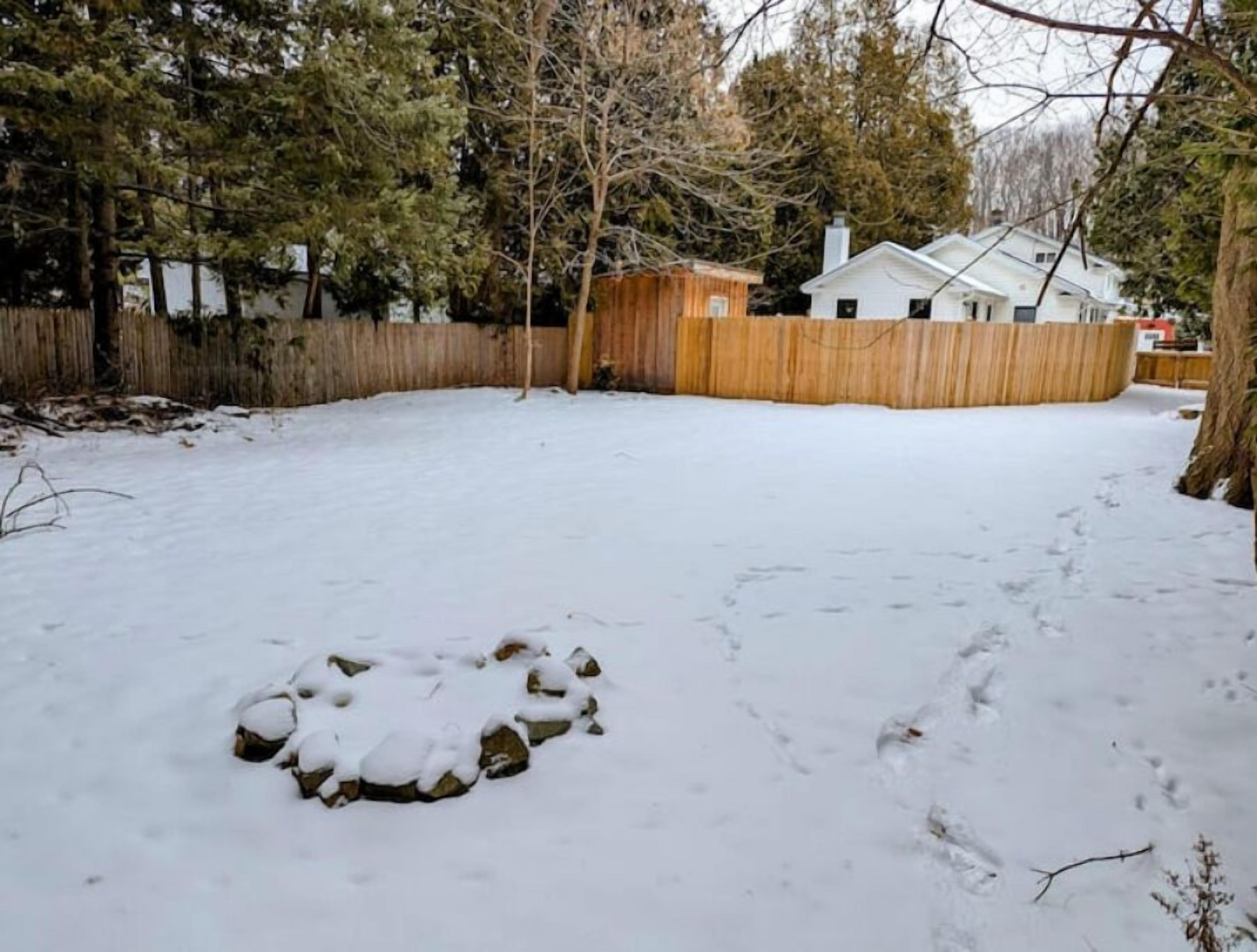A snow-covered backyard with a wooden fence, a house in the background, and a tree in the foreground.