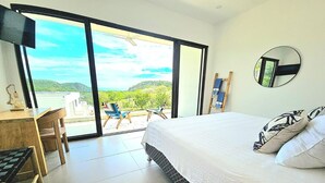 Een slaapkamer met een bed, een bureau en een stoel. Er is een groot raam met uitzicht op groen en heuvels.