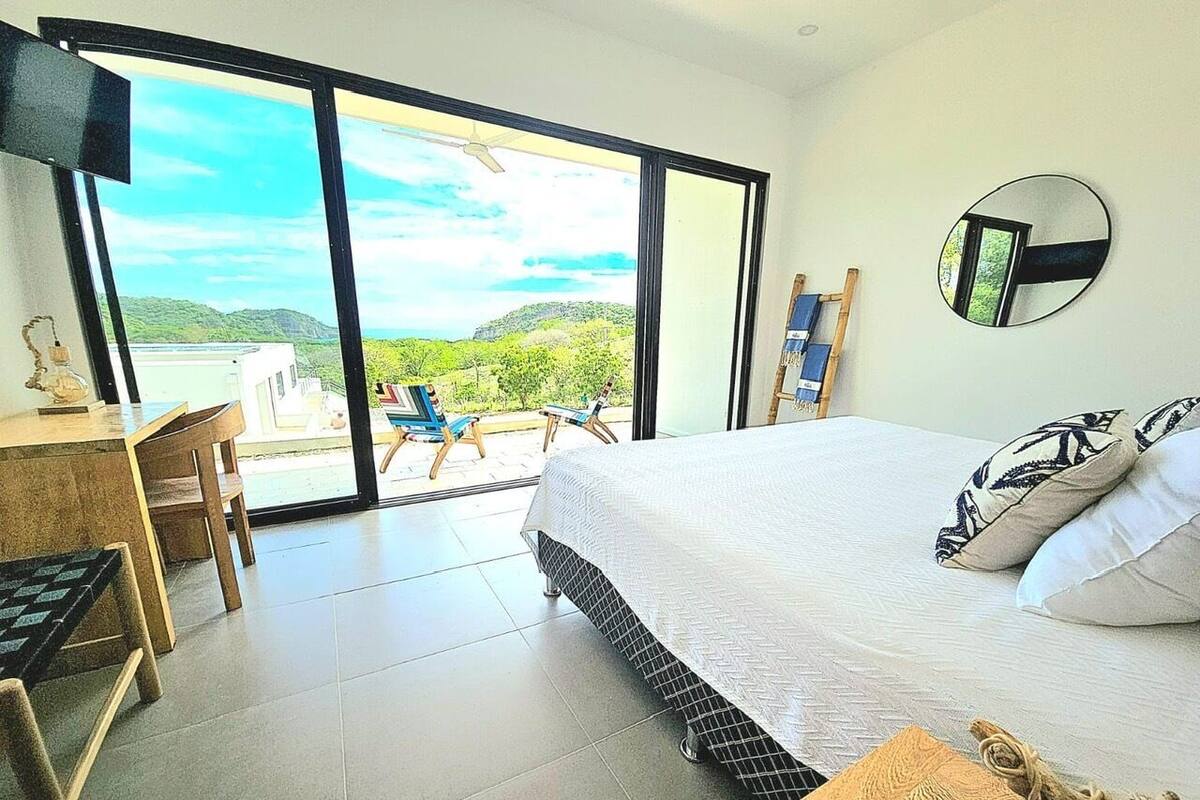 A bedroom with a bed, a desk, and a chair. There is a large window with a view of greenery and hills.