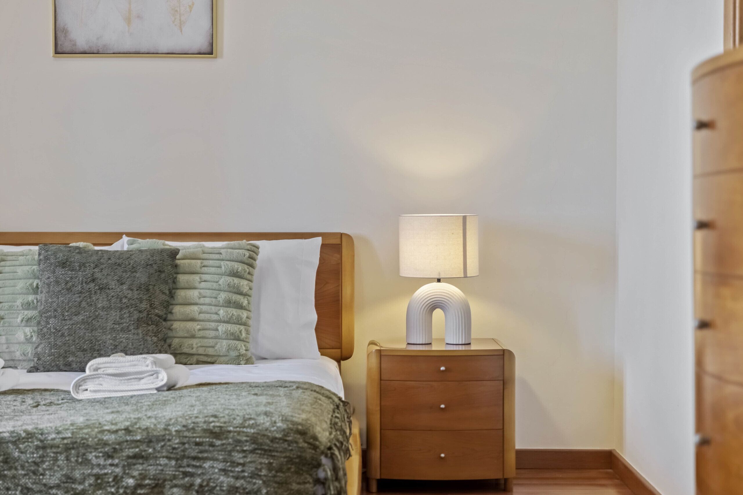 Cama bem arrumada com cabeceira de madeira, abajur e pequena mesa de cabeceira com gavetas.