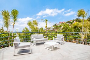 An outdoor patio with white furniture, surrounded by palm trees and a view of a hillside with buildings. - Amendoeira Residence by Homie (Ponta do Sol)