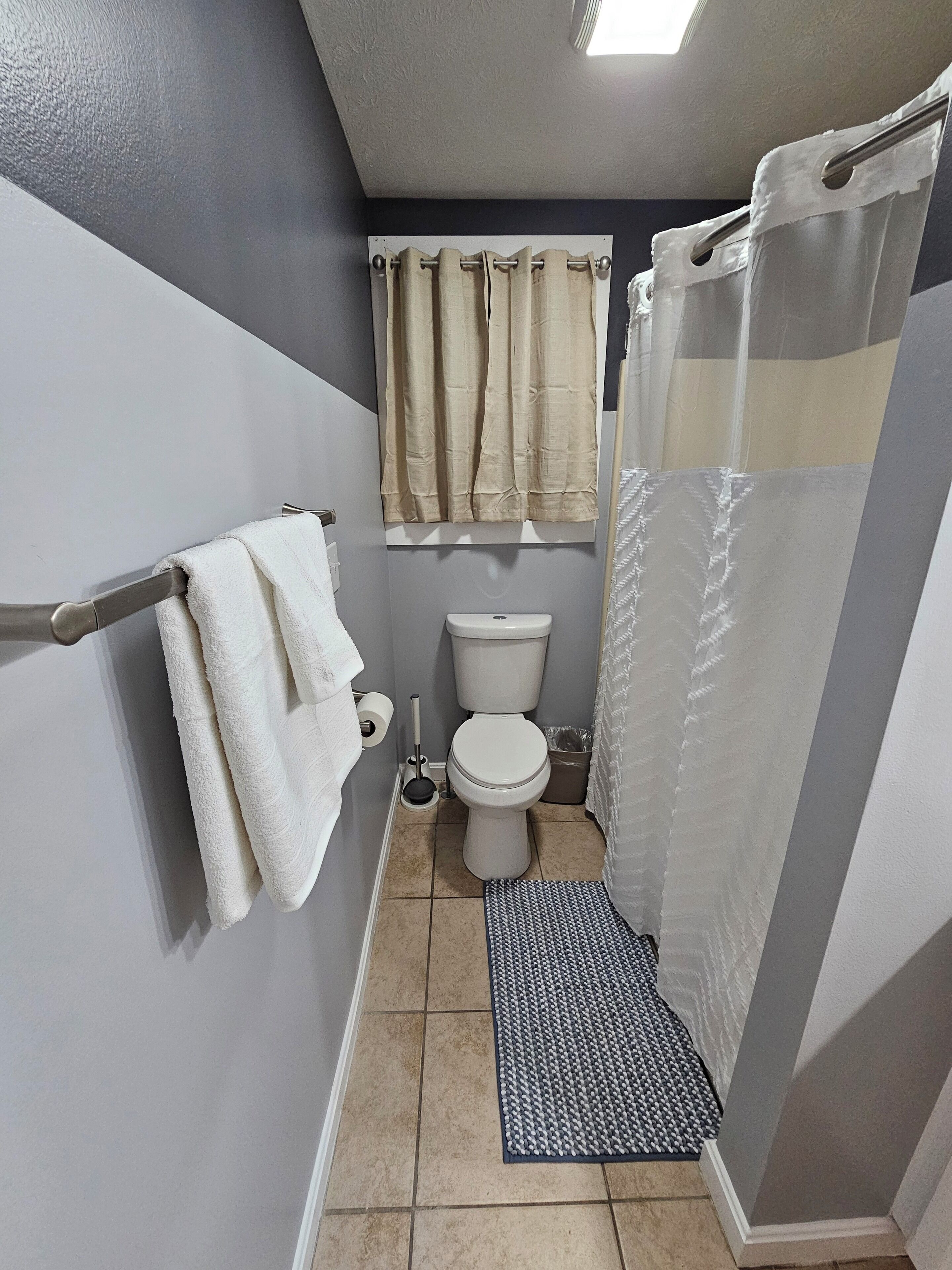 Een badkamer met toilet, douchegordijn, handdoekrek met handdoeken, toiletpapierhouder en een vloermat.