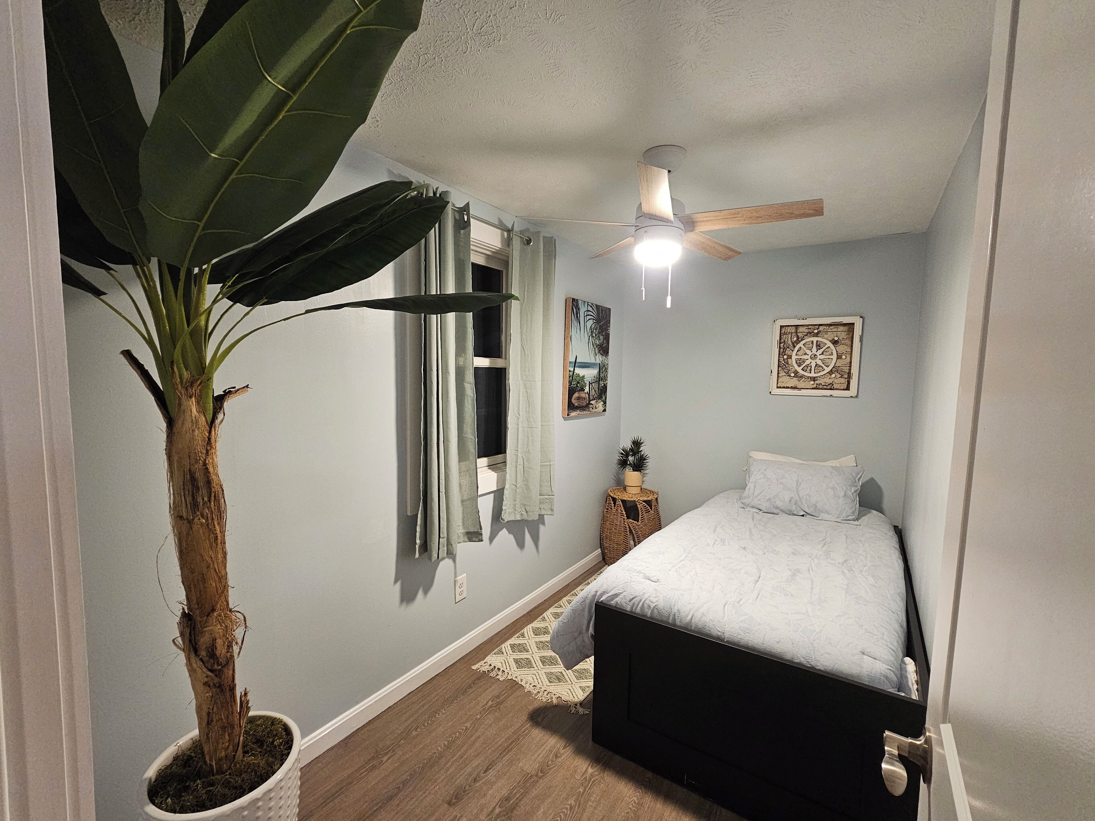 A bedroom with a bed, a ceiling fan, a window with curtains, a potted plant, and wall art.