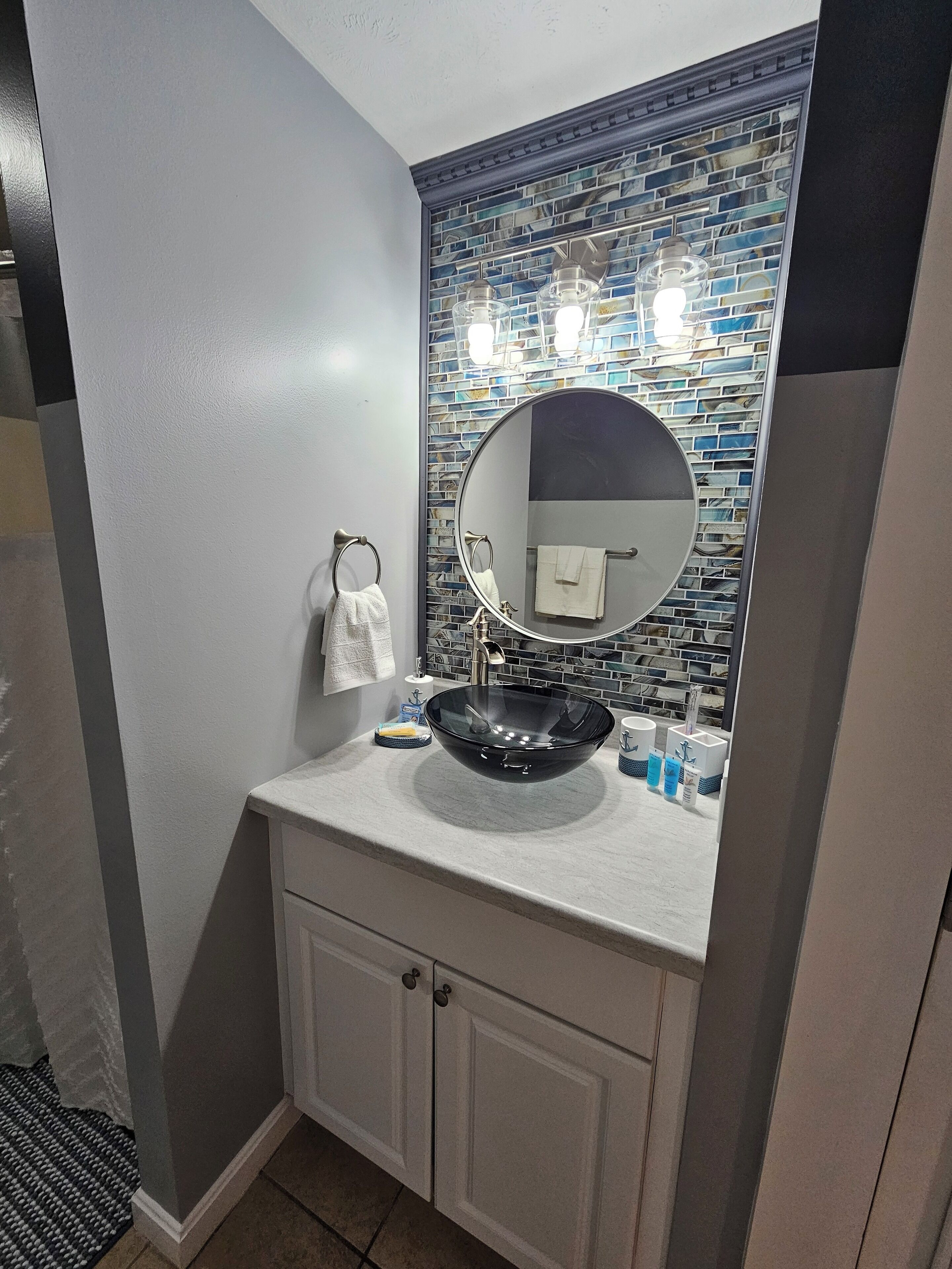 Un baño con un espejo redondo, un lavabo de vidrio y un salpicadero de azulejos.