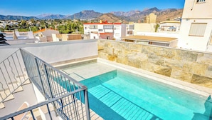Une piscine sur le toit, bordée d’un mur en pierre et d’une rambarde en métal, offrant une vue sur un quartier résidentiel avec des montagnes en arrière-plan.