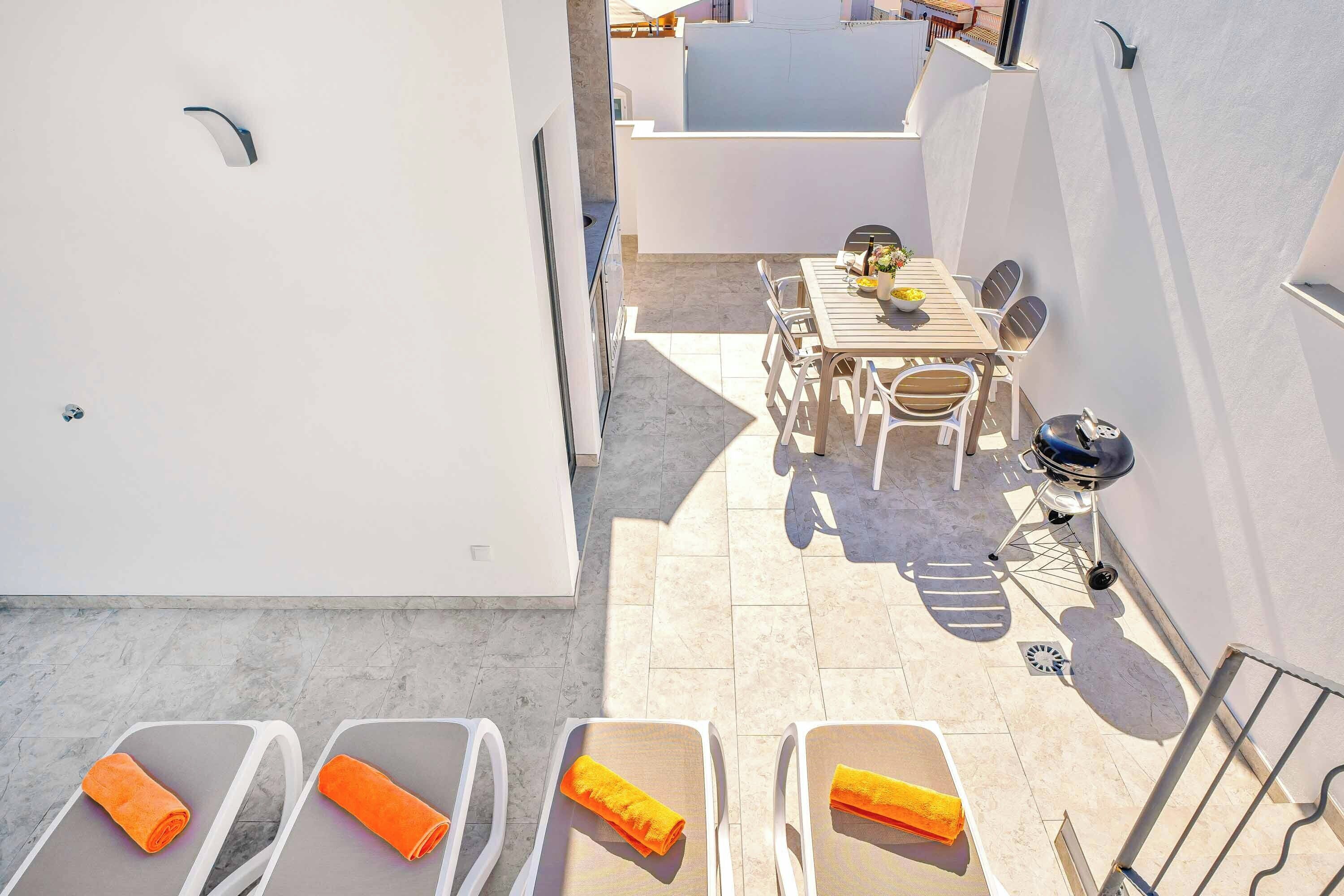Eine Dachterrasse mit Tisch, Stühlen, einem Grill und Liegestühlen mit Handtüchern.