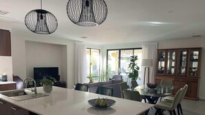A modern kitchen with a central island, a dining area with a glass table, and a view of the outdoors. - Mid-century Elegance in a leafy garden (Padbury)