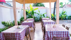 Une terrasse extérieure avec des tables et des chaises, recouverte d’un toit en bois, entourée de plantes et d’un mur blanc.