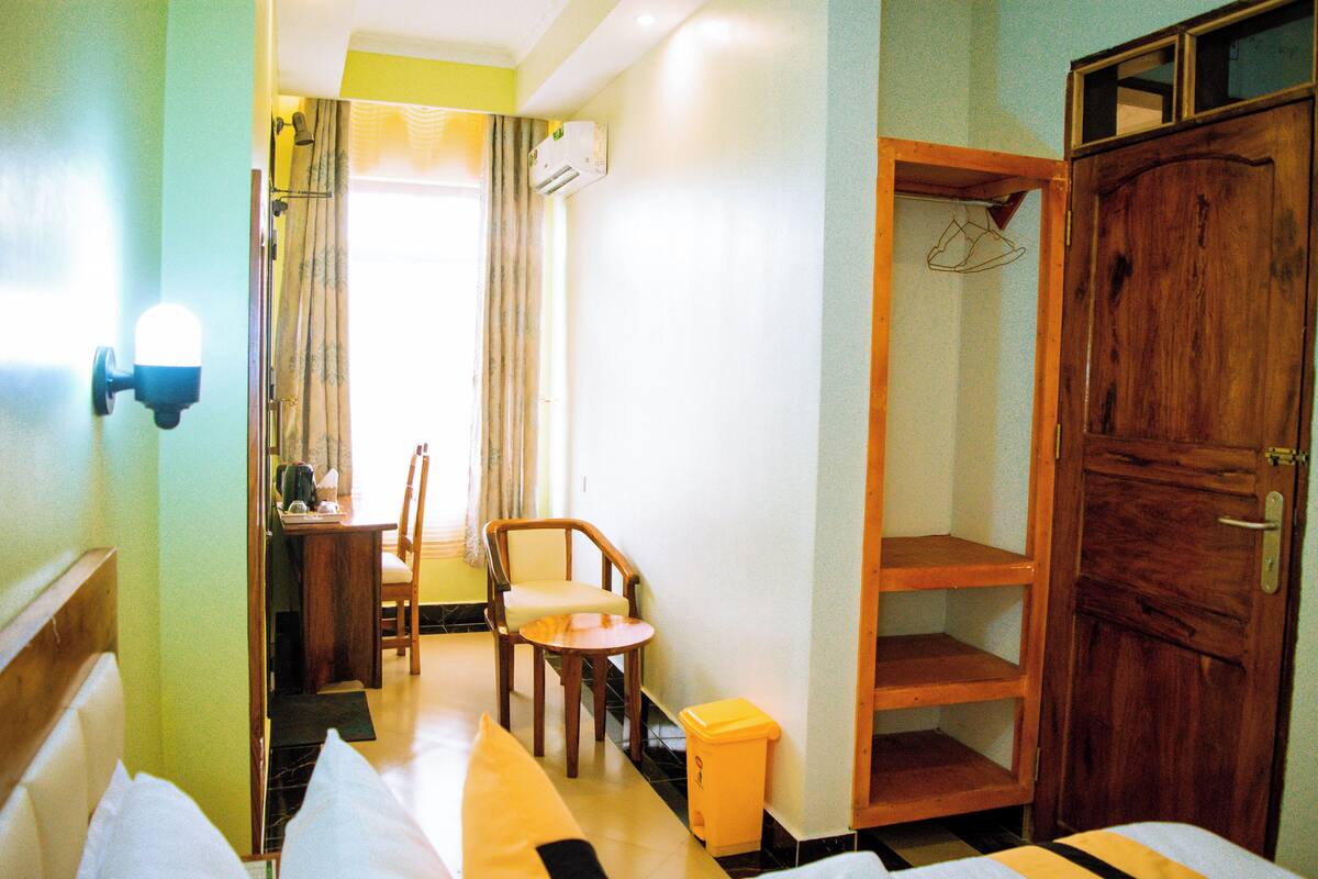 A hotel room with a bed, a small table, a chair, a wooden wardrobe, and a bathroom visible through an open door.